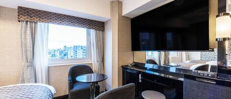 Habitación de hotel con cama, dos sillas, una mesita, televisor y ventana con vista a la ciudad.