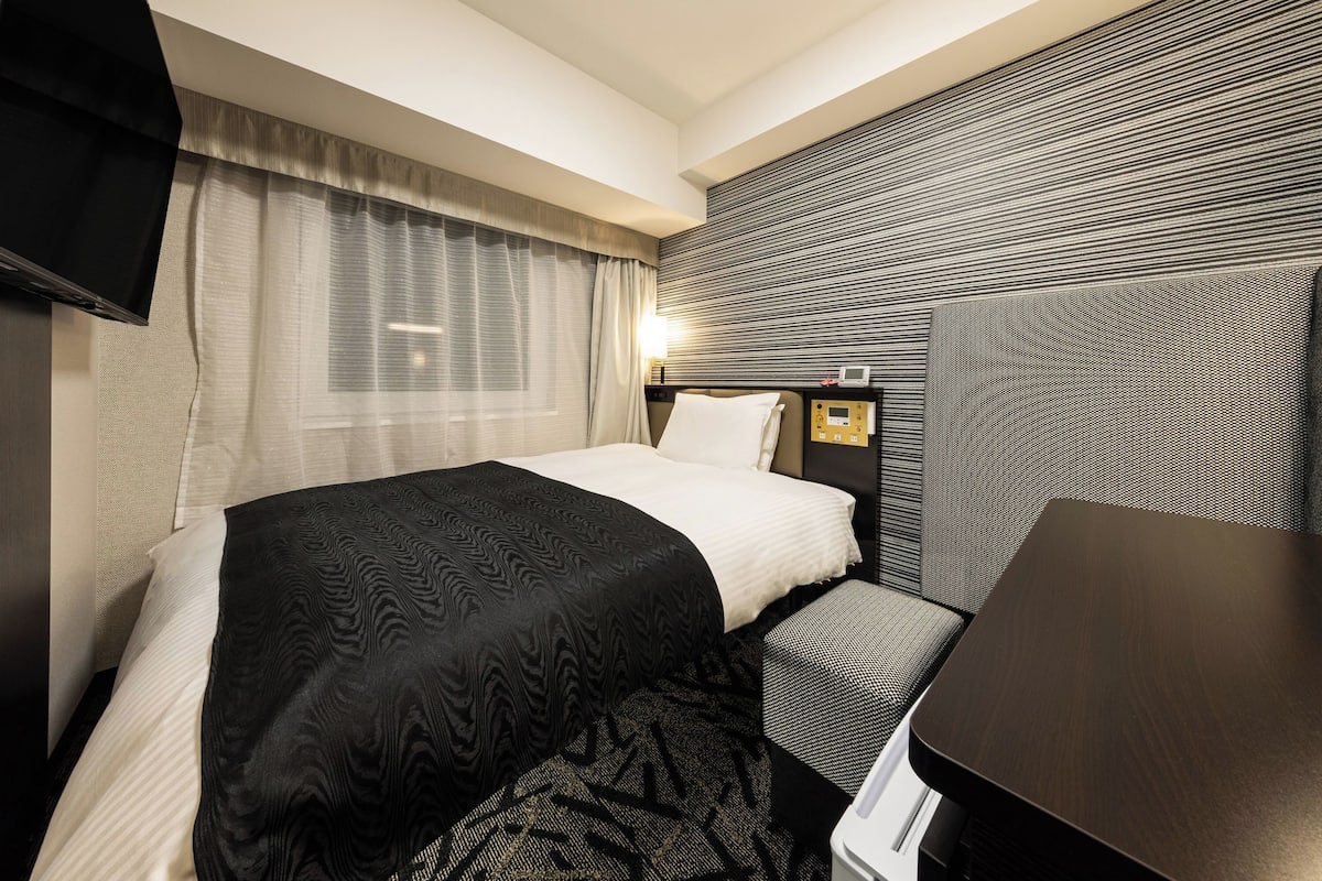A hotel room with a bed, a desk, a TV, and a wall-mounted control panel.