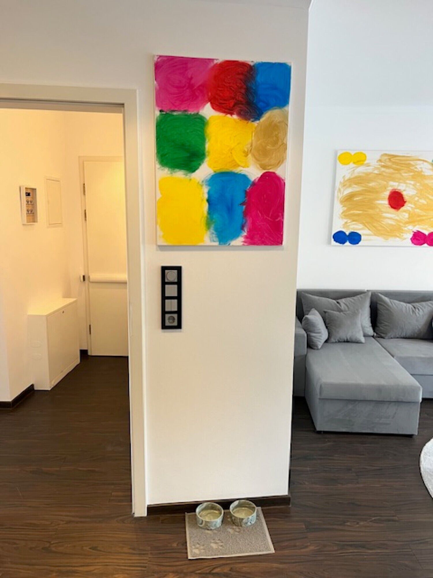 Ein modernes Wohnzimmer mit einem grauen Sofa, einem farbenfrohen abstrakten Gemälde und einer Tür mit Lichtschalter.