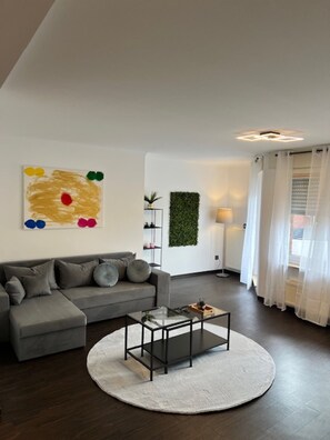 En moderne stue med en grå sofa, et glas sofabord, et rundt gulvtæppe, et kunstværk på væggen, grøn vægdekoration og et vindue med lette gardiner.