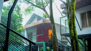 A modern building with a staircase, a tree with moss, and a view of a red building in the background. - Lakkidi Mist Resort (Vythiri)