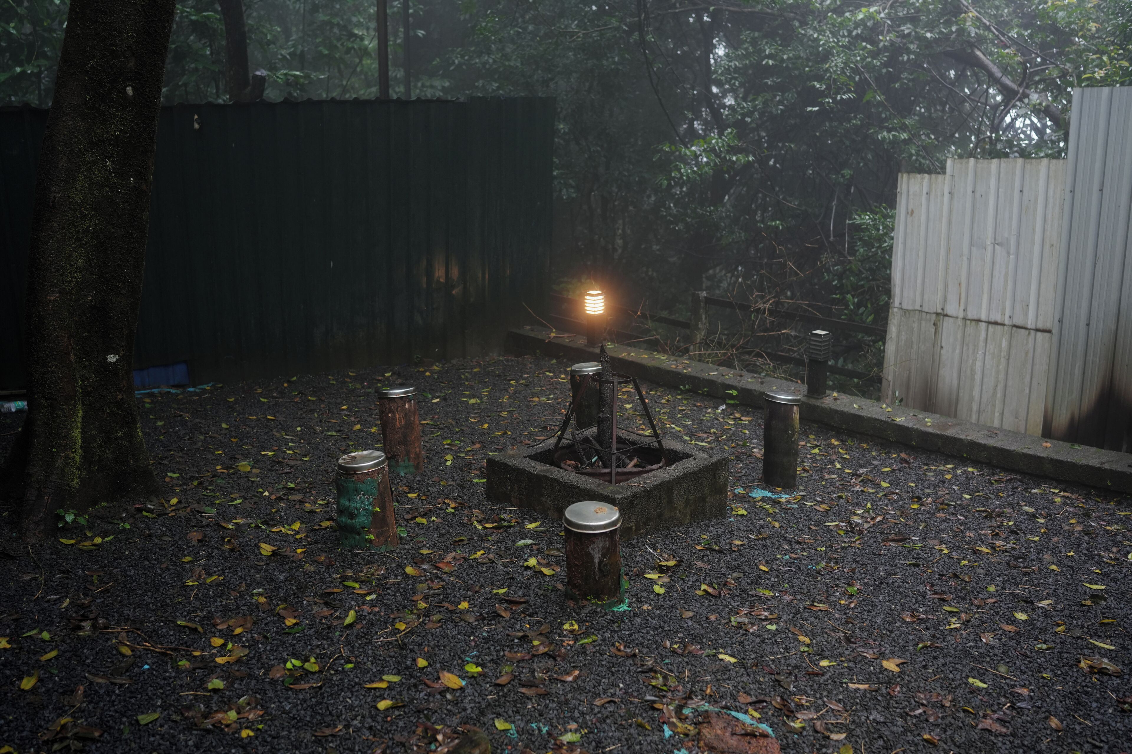 A forested area with a metal structure, possibly a fire pit, surrounded by scattered cans and leaves.