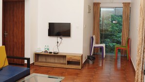 A modern living room with a flat-screen TV mounted on the wall, a glass coffee table, and a wooden bench. There are two chairs, one purple and one green, and a wooden door on the left. - Lakkidi Mist Resort (Vythiri)