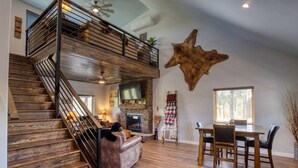 A spacious living area with a wooden staircase, a fireplace, a dining table, and a large wall-mounted animal hide. - Cabin Retreat in the White Mountains of Arizona (Show Low)