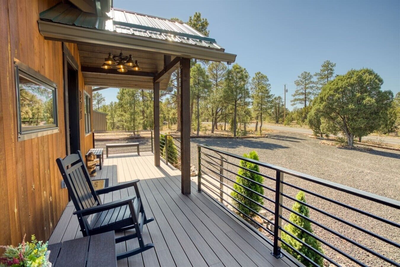 A wooden deck with a bench and a table, a covered area with hanging lights, and a view of a gravel area with trees.