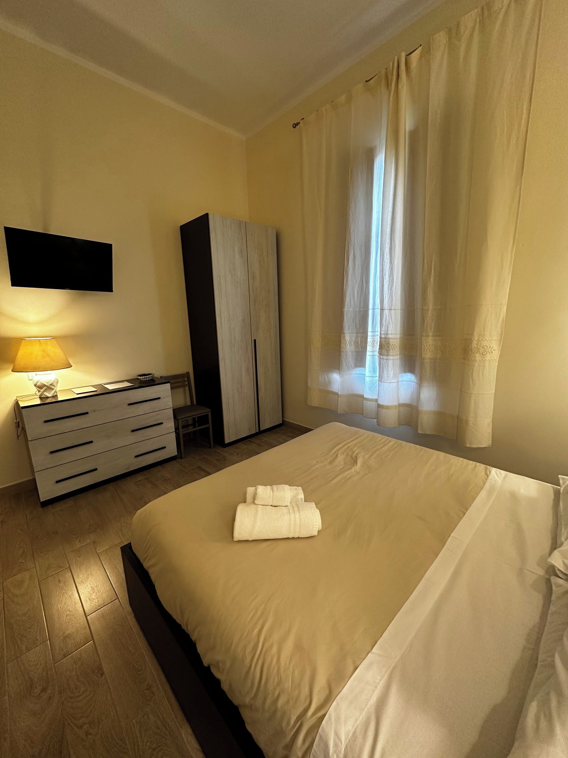 Una habitación de hotel con cama, escritorio con lámpara, armario y televisor en la pared.