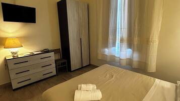 Una habitación de hotel con cama, escritorio con lámpara, armario y televisor en la pared.