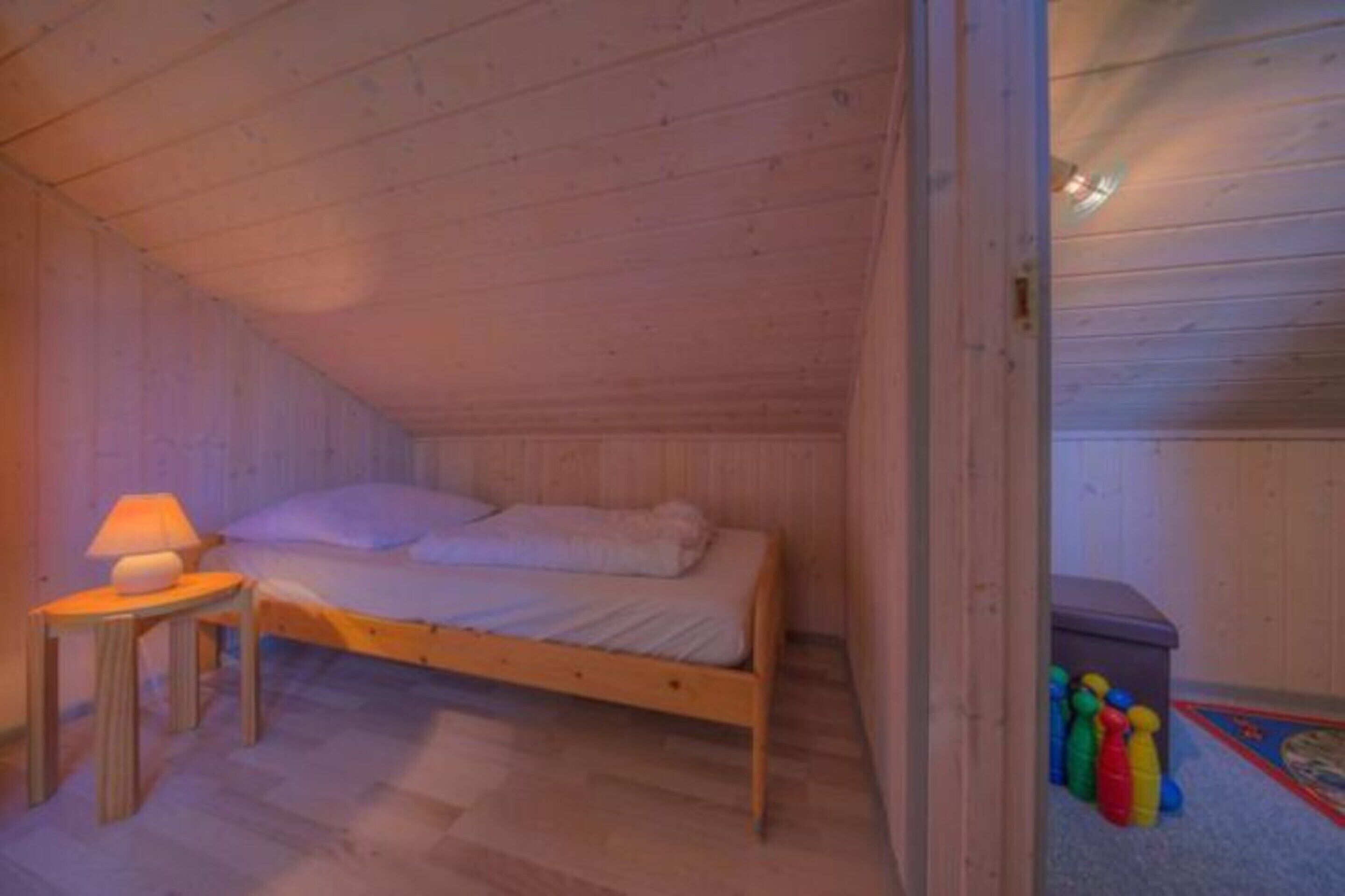 Ein kleines Zimmer mit einem Einzelbett, Holzboden, Nachttisch mit Lampe und einer kleinen Aufbewahrungsbox mit bunten Spielsachen.