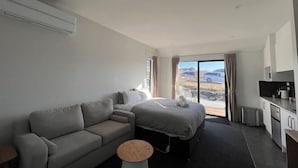 Een moderne hotelkamer met een bank, een bed, een klein rond tafeltje, een kitchenette en een groot raam met uitzicht.