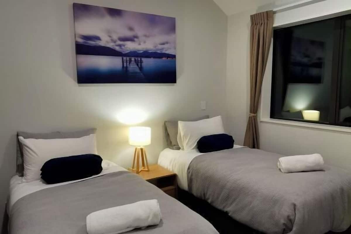 Une chambre d’hôtel avec deux lits, une table de chevet avec une lampe, une fenêtre avec des rideaux et un tableau au mur.