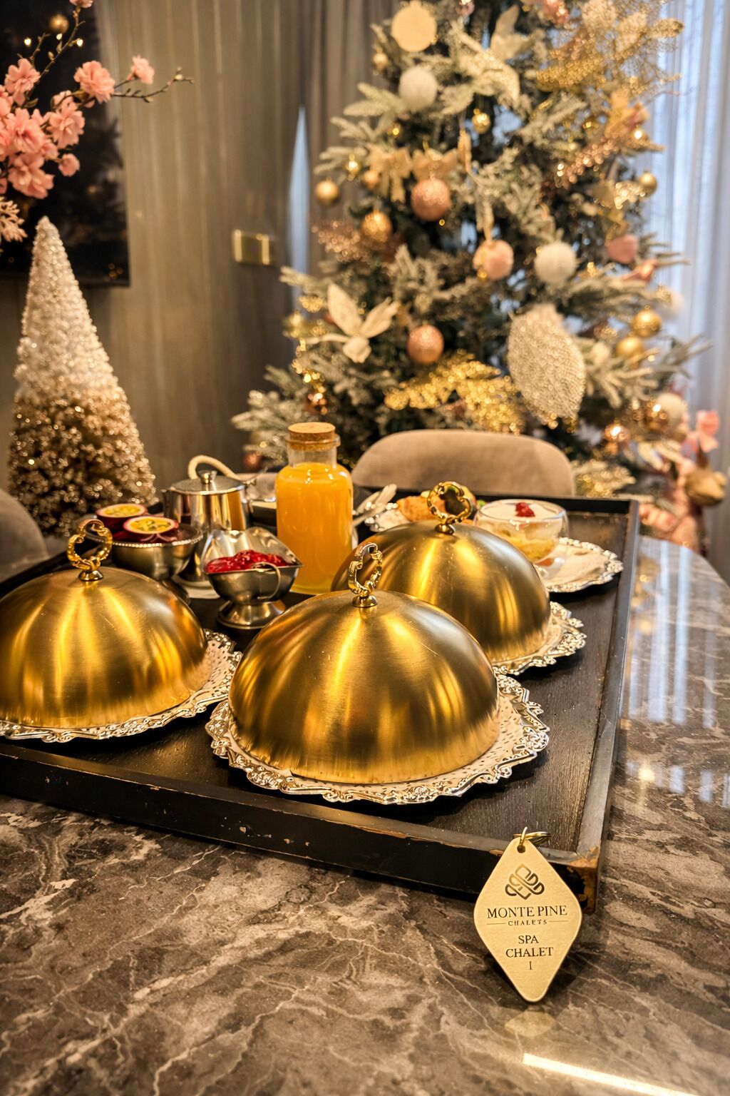 Un plateau avec des couvercles dorés en forme de dôme, une bouteille de jus d’orange et une étiquette indiquant « Monte Pino Chalet Spa Chalet I ».