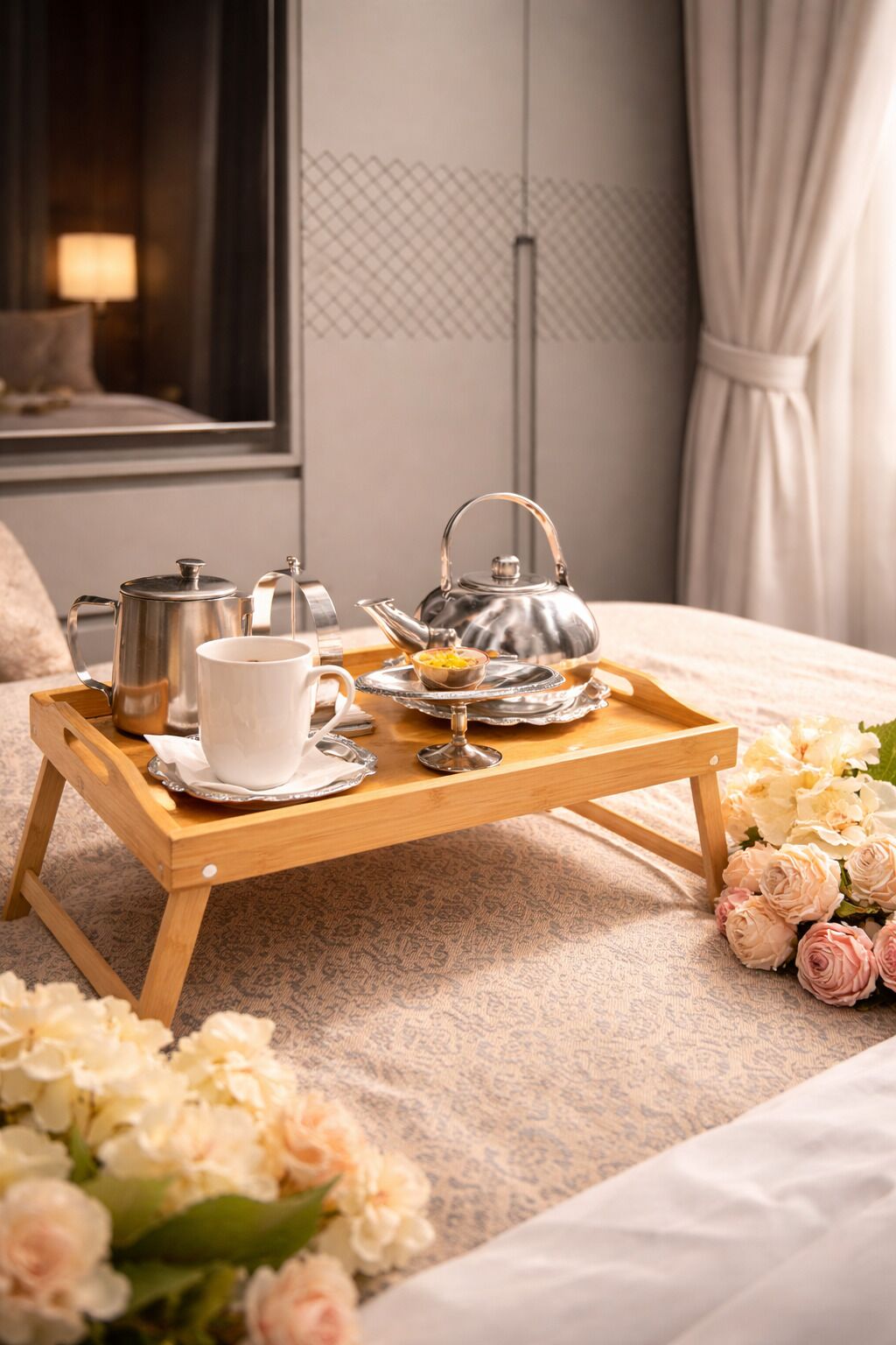 A wooden tray with a teapot, a cup, and a small bowl on a bed with a floral arrangement.