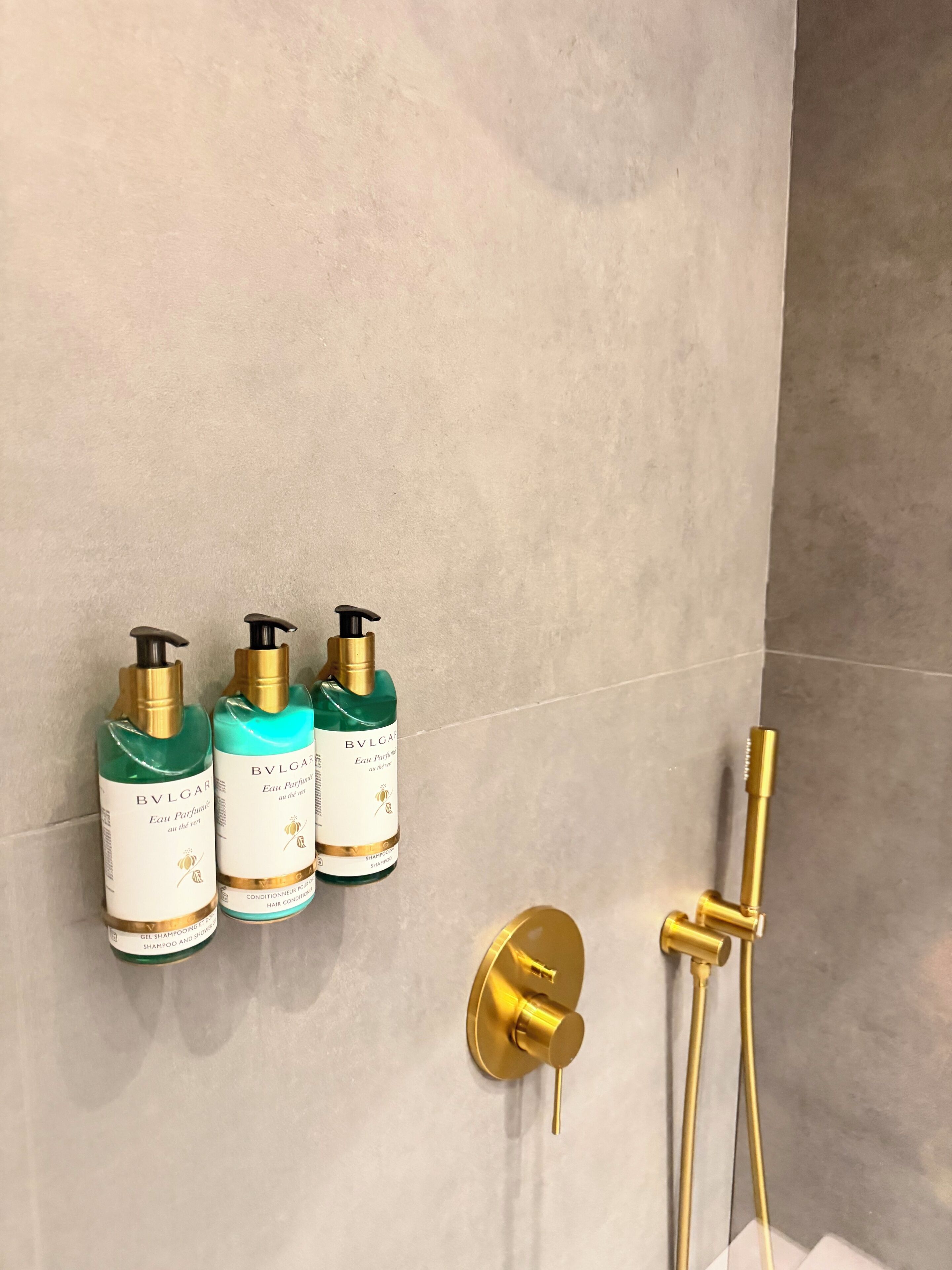 Une salle de bain avec une pomme de douche dorée et trois flacons de parfum Bvlgari Eau de Parfum accrochés au mur.