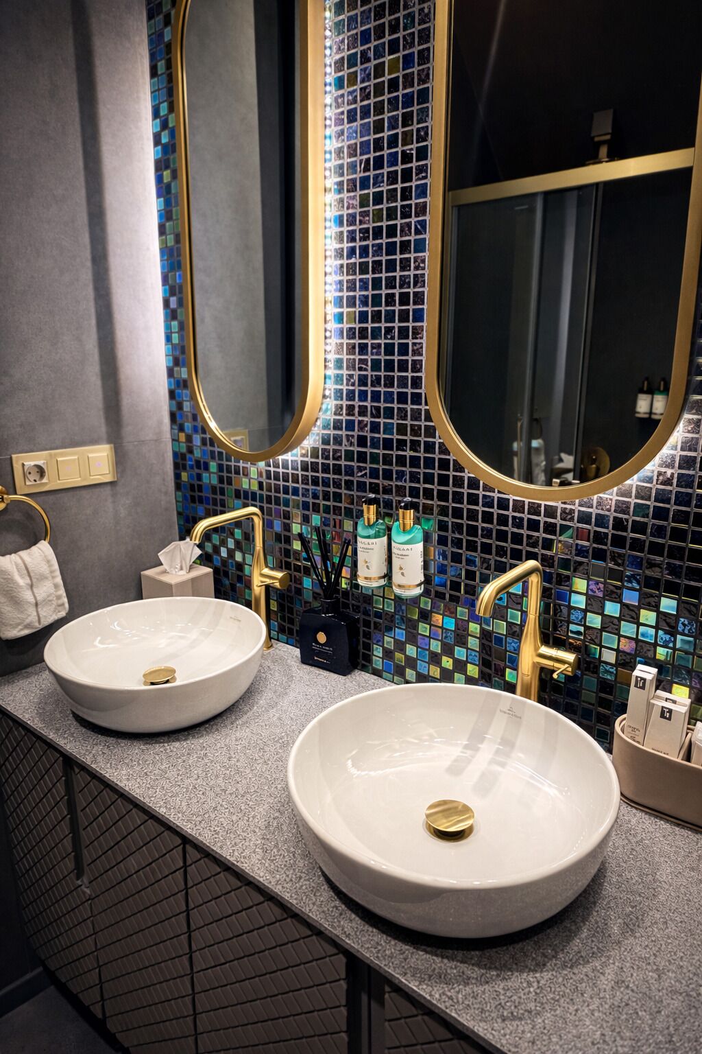 A bathroom with two round white sinks, gold faucets, and a mosaic tile backsplash.
