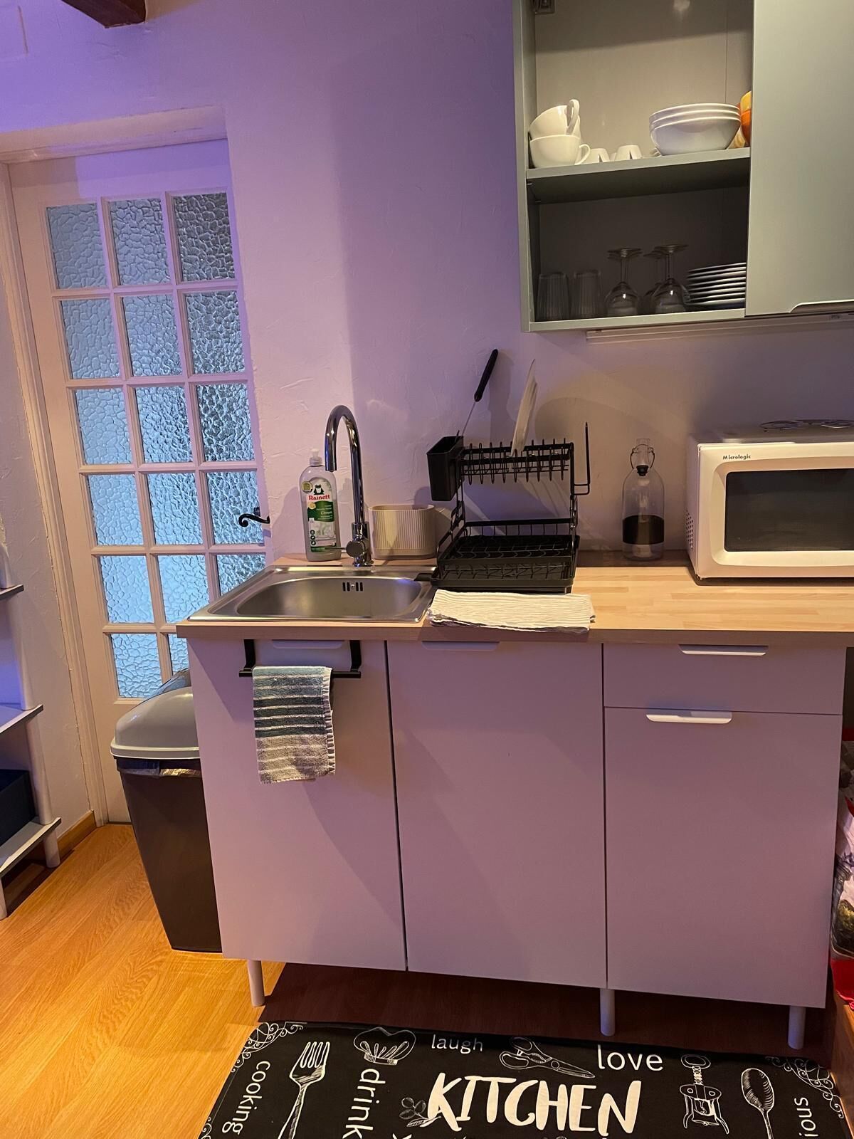 Una cucina compatta con lavello, forno a microonde ed espositore per piatti. Si nota una finestra con vetro decorato e un asciugamano appeso all'armadietto.