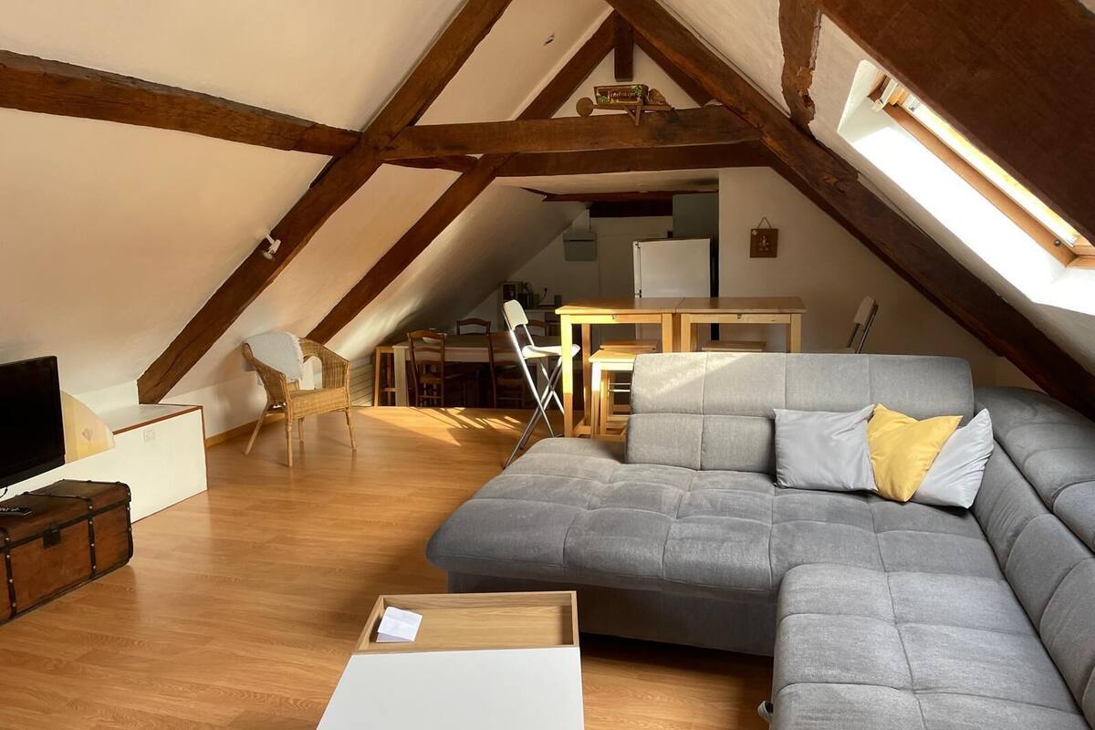 Ein Loft-Wohnraum mit geneigter Decke, Holzbalken, einem grauen Ecksofa, einem Essbereich mit Tisch und Stühlen sowie einer kleinen Küchenzeile im Hintergrund.