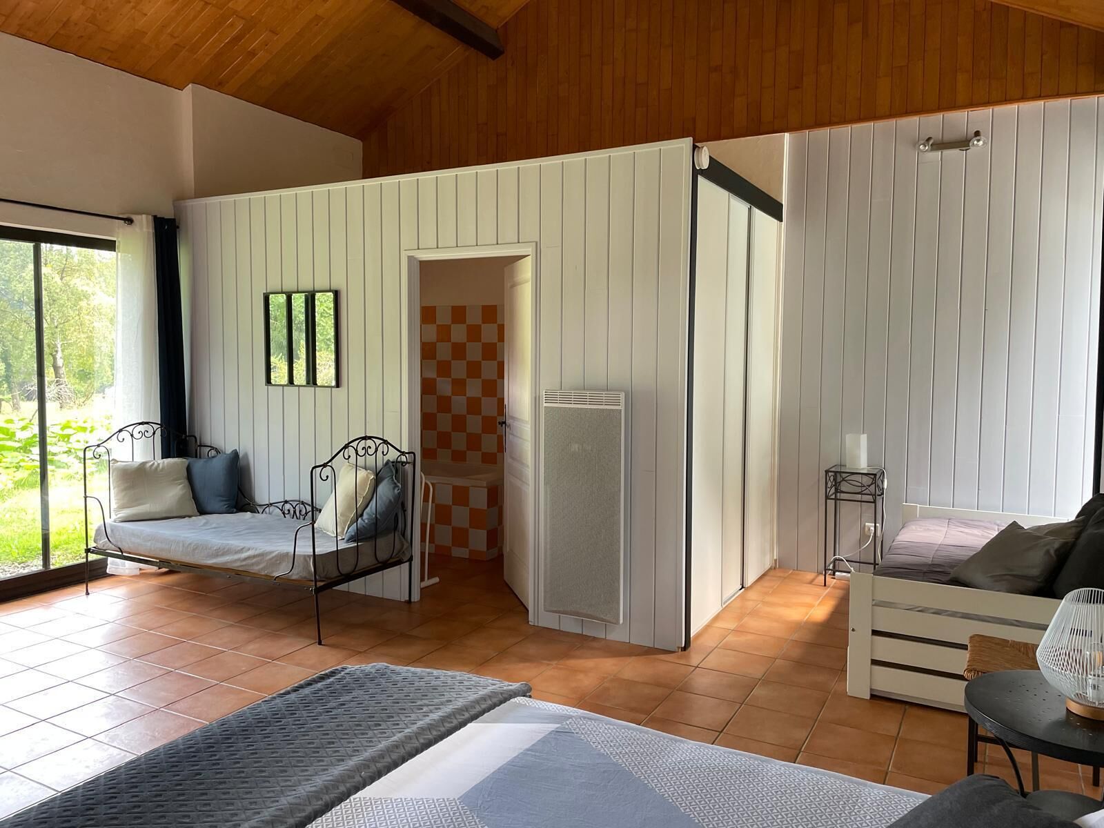 Ein Zimmer mit einem Bett, einem kleinen Tisch und einem Stuhl. Es gibt ein Fenster mit Vorhängen, eine Tür und eine karierte Wand.