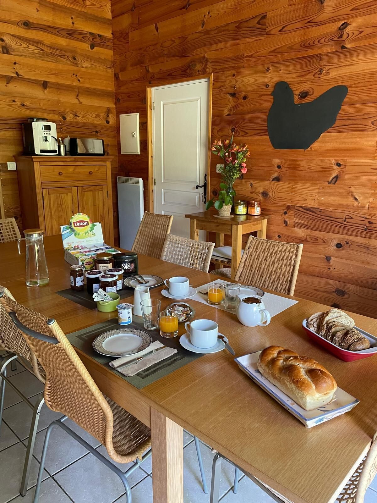 A wooden dining table set for breakfast with wicker chairs, a coffee maker, Lipton tea, and a variety of breakfast items.