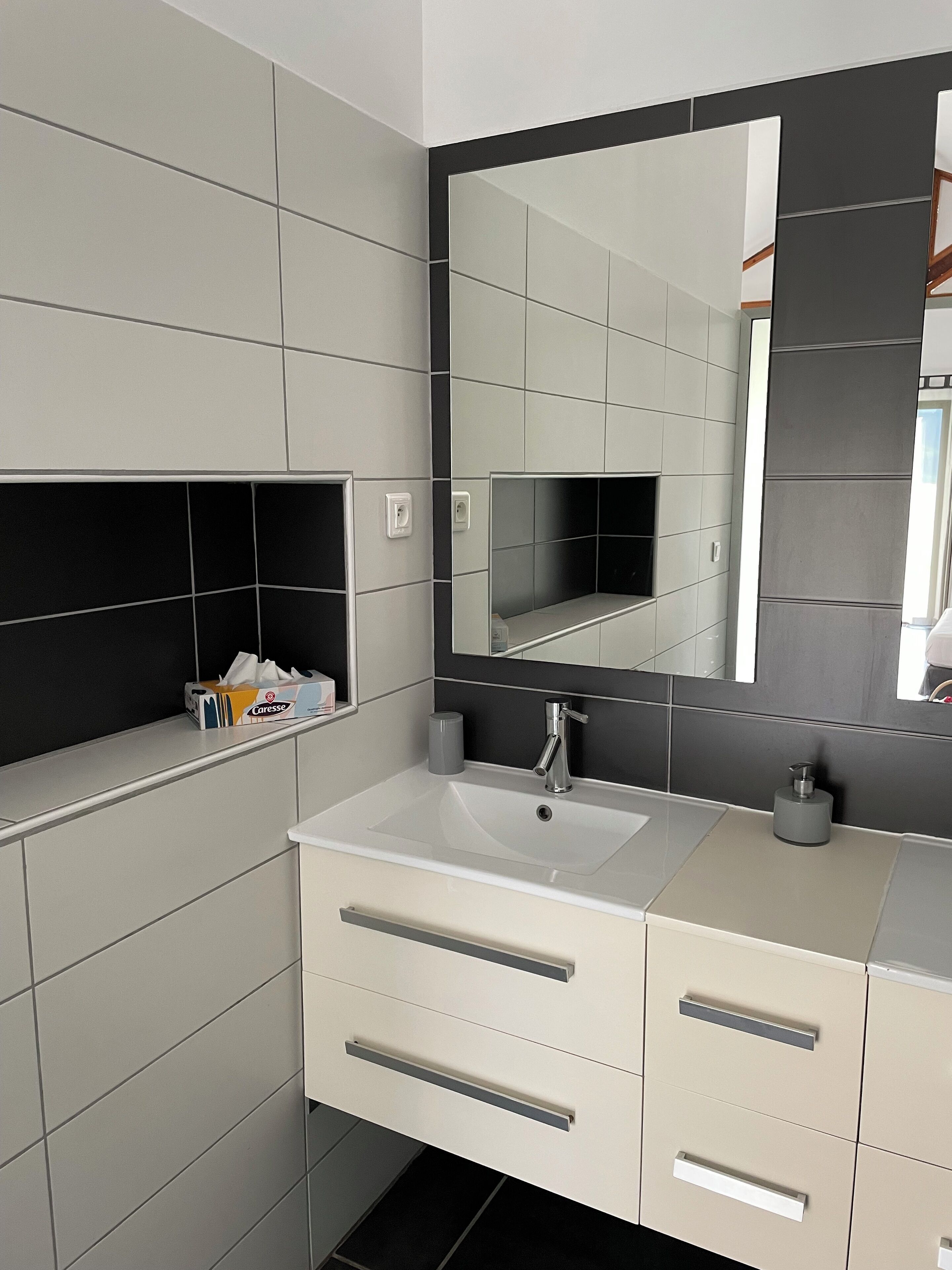 Un bagno moderno con un ampio specchio, piastrelle bianche e grigie, un lavandino e un mobile contenitore con cassetti.