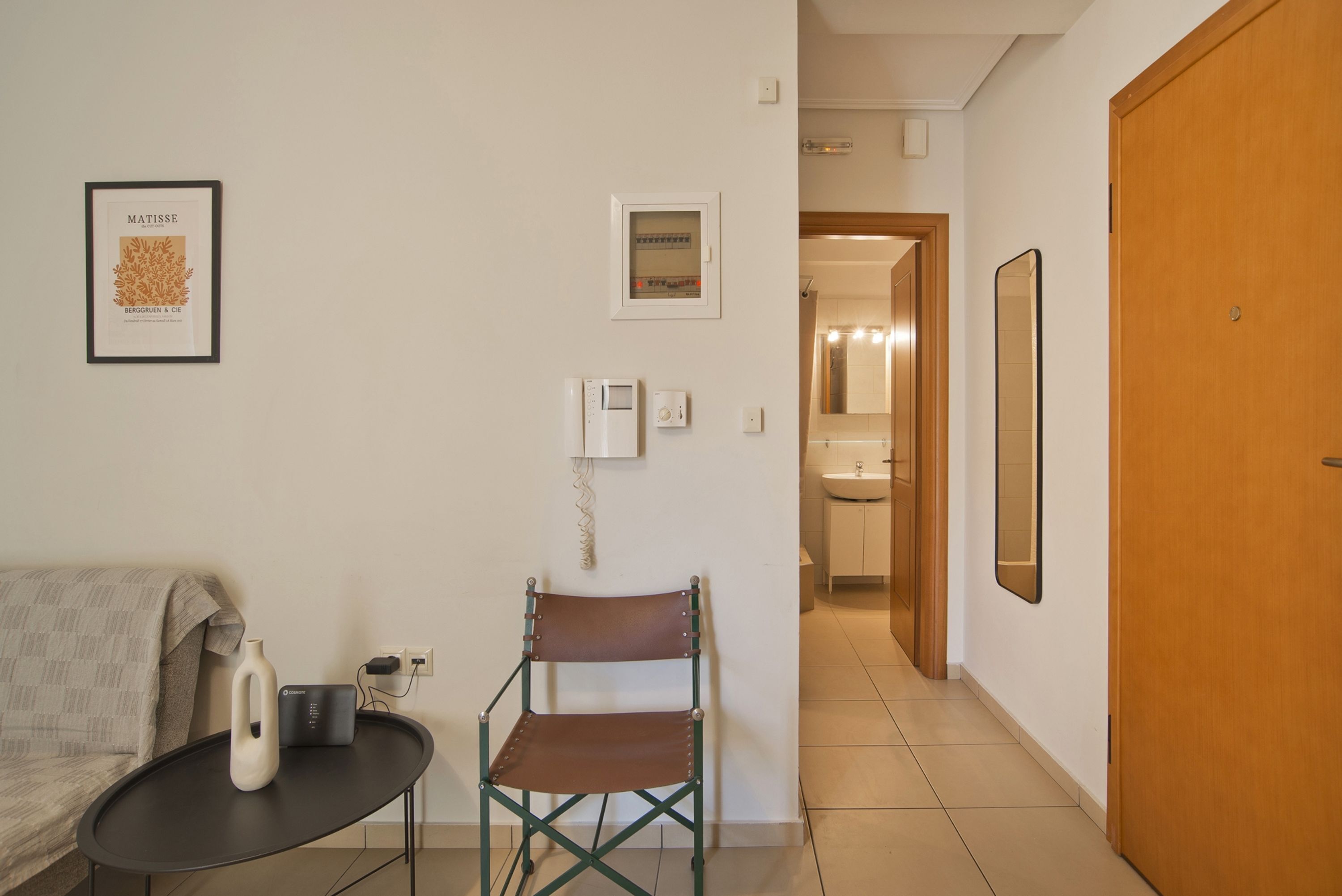 A hotel room with a sofa, a small table with a bottle and a power strip, a folding chair, a framed artwork, a thermostat, a mirror, and a wooden door.
