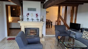 Un salon avec une cheminée, deux fauteuils, une table basse et un téléviseur. Au fond de la pièce, on aperçoit un coin repas avec une table et des chaises.