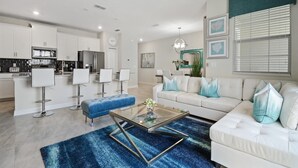 A modern living room with a white sofa, blue ottoman, and a glass coffee table. There is a kitchen area with white cabinets and a dining area with white chairs.