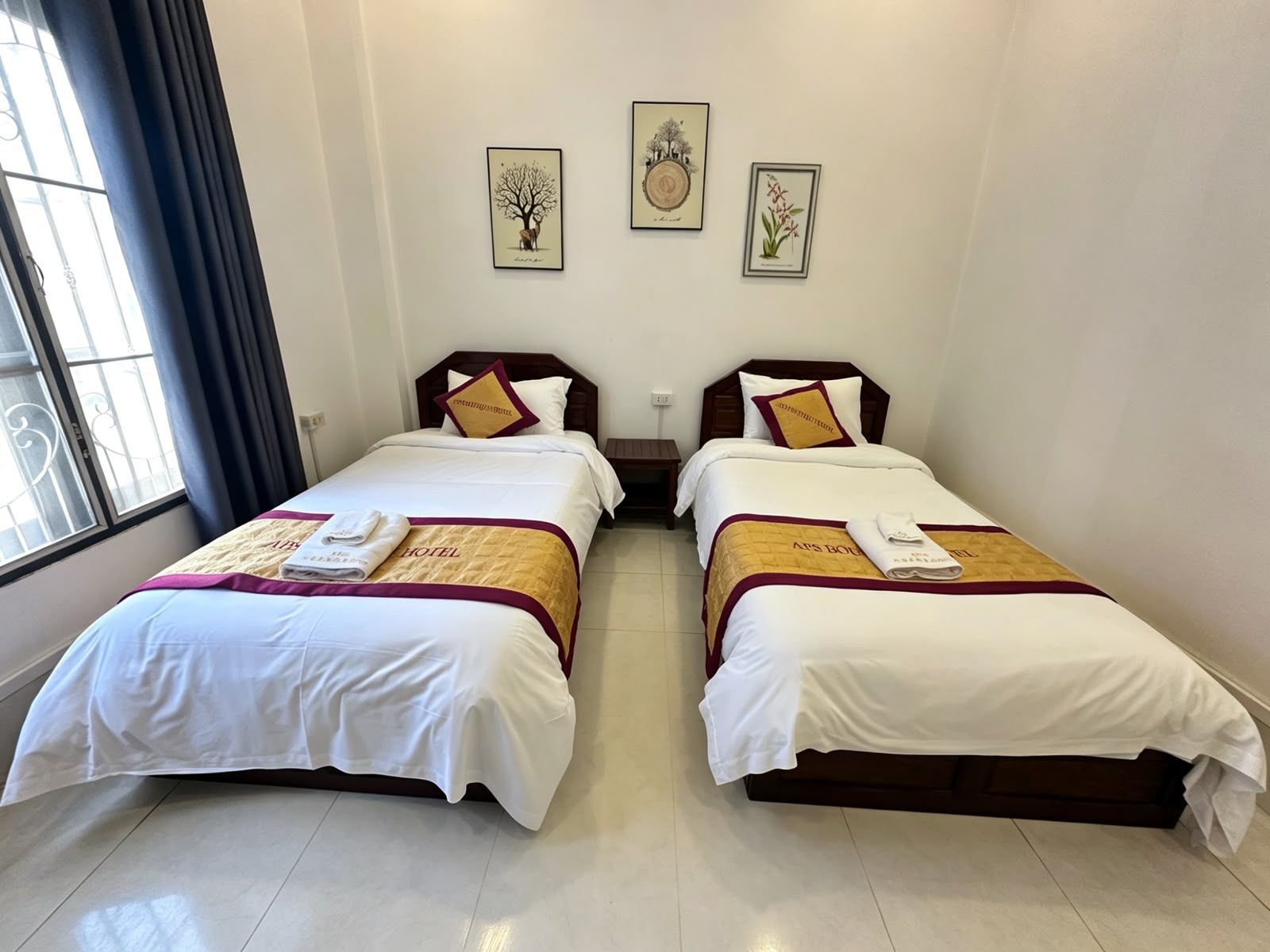 A hotel room with two beds, each with a white bedspread and a maroon accent. There are framed botanical prints on the wall above the beds.