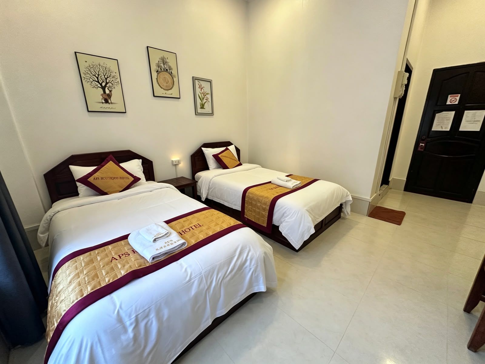 A hotel room with two beds, each with a white and maroon bedspread, and a small nightstand between them. There are framed botanical prints on the wall above the beds.