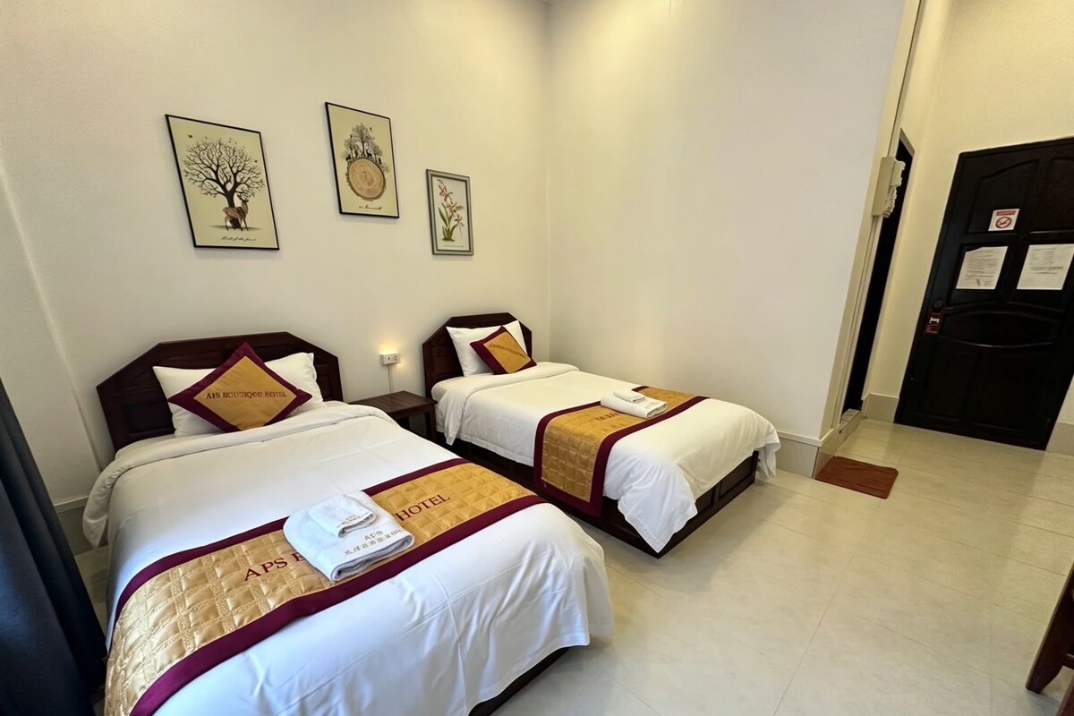 A hotel room with two beds, each with a white and maroon bedspread, and a small nightstand between them. There are framed botanical prints on the wall above the beds.