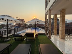 A rooftop pool area with lounge chairs and umbrellas, surrounded by stone pillars and a view of the sunset. - A P S Boutique Hotel (Vang Vieng)