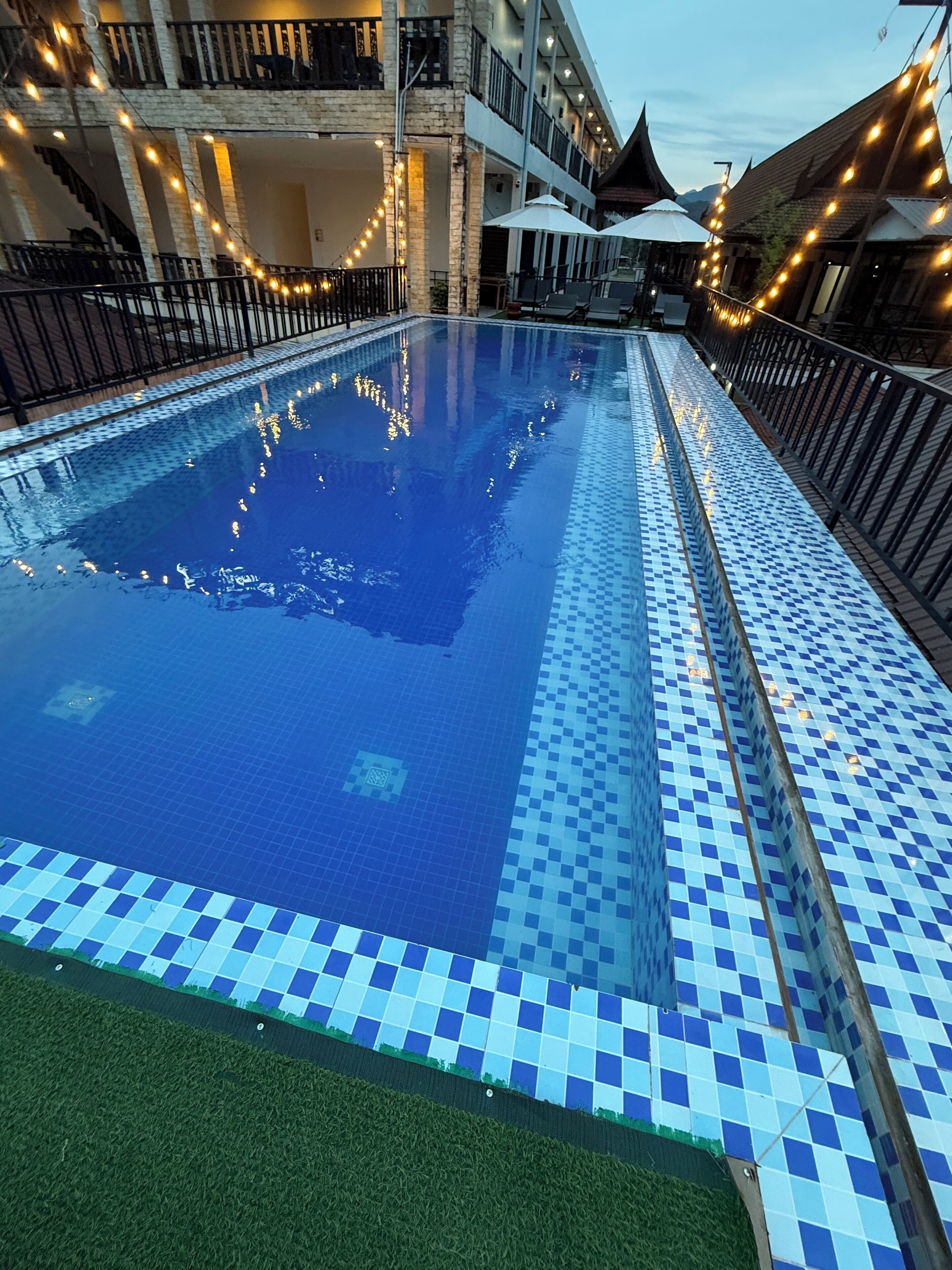A swimming pool with a checkered tile border, surrounded by a railing and string lights.