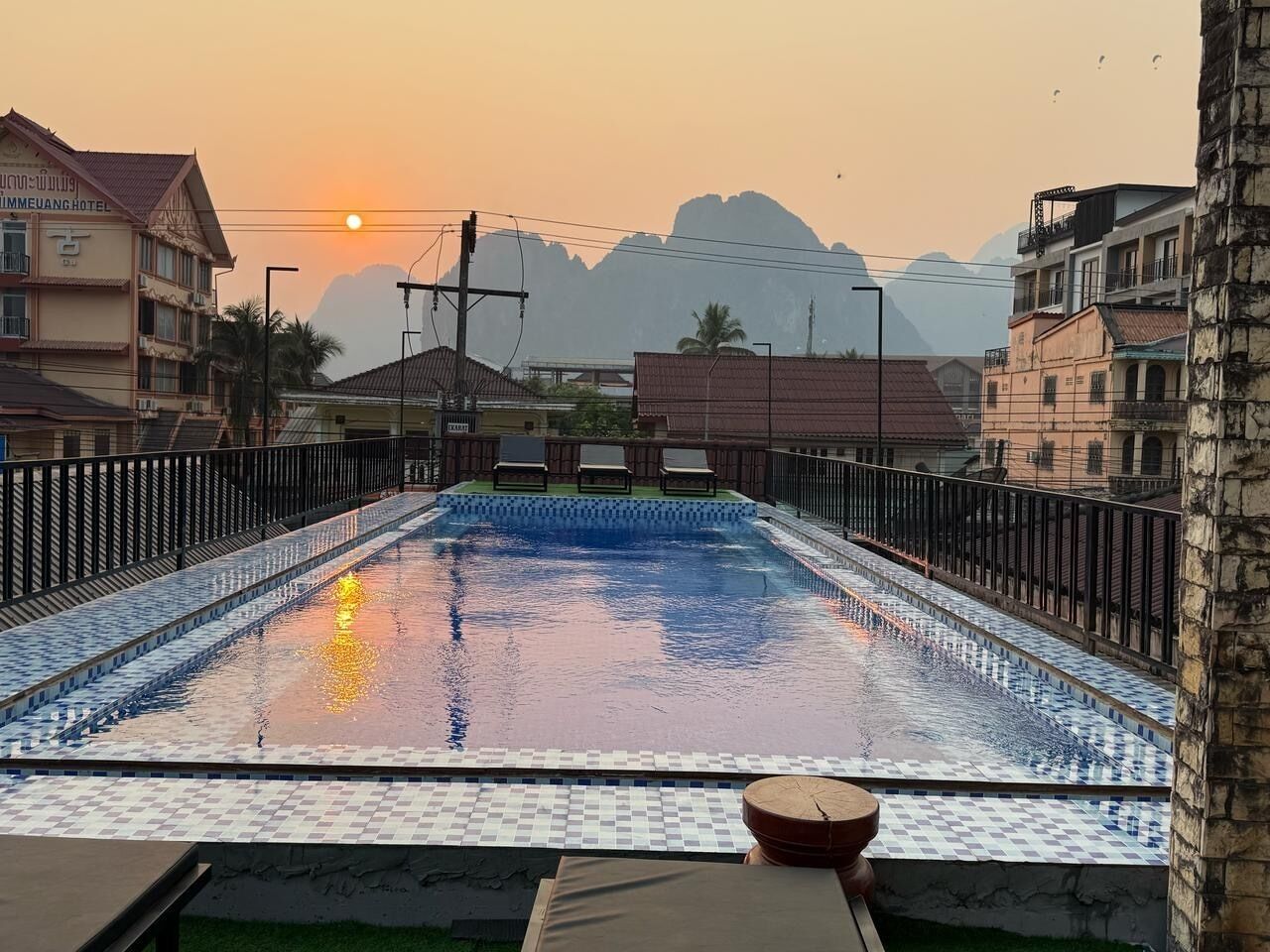 En swimmingpool med ett kaklat poolområde, omgiven av ett staket, och en solnedgång i bakgrunden.