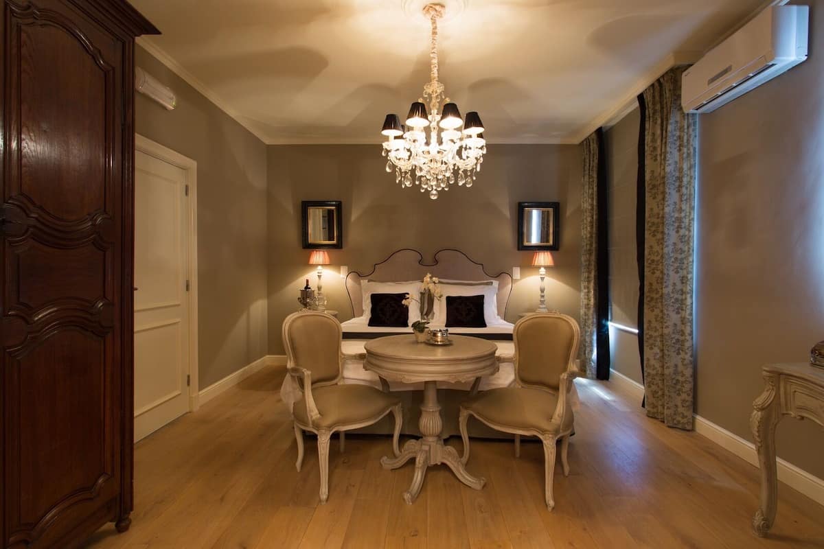 A room with a chandelier, a bed with a headboard, two chairs, a table, and two wall-mounted lamps.