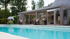 A modern poolside area with a glass-enclosed lounge, outdoor seating, and a clear view of the pool. - Villa Reynaert (Maaseik)