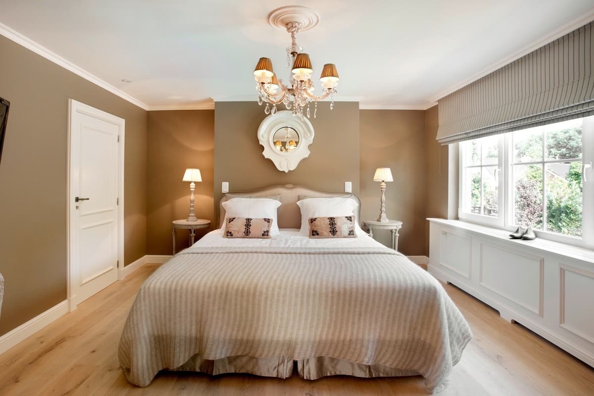 A bedroom with a large bed, two bedside tables with lamps, a chandelier, a mirror above the bed, and a window with a view of greenery.