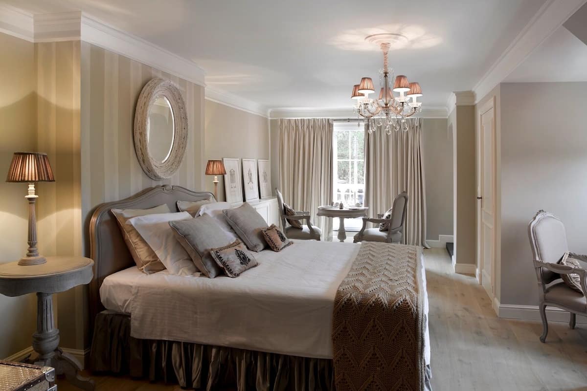 A spacious bedroom with a large bed, a chandelier, a round mirror, and a dining table with chairs.