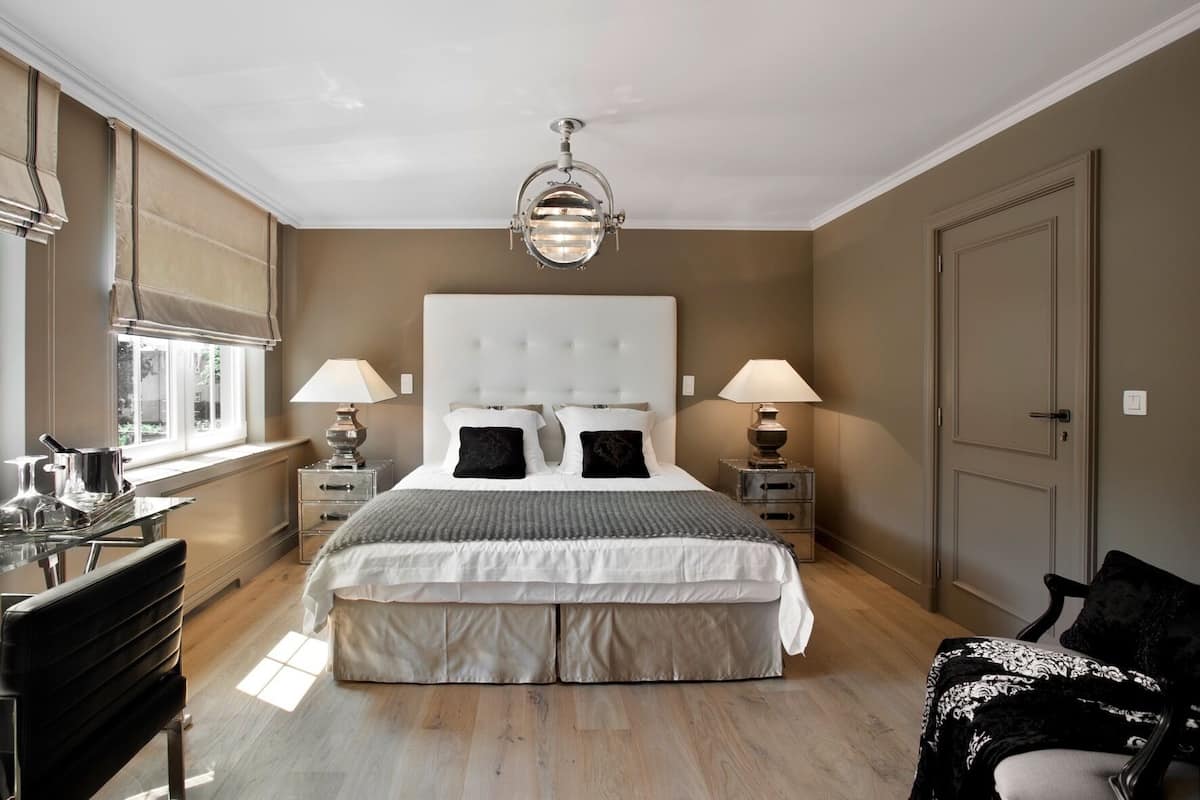 A bedroom with a large bed, two bedside tables with lamps, a desk with a chair, and a window with blinds.