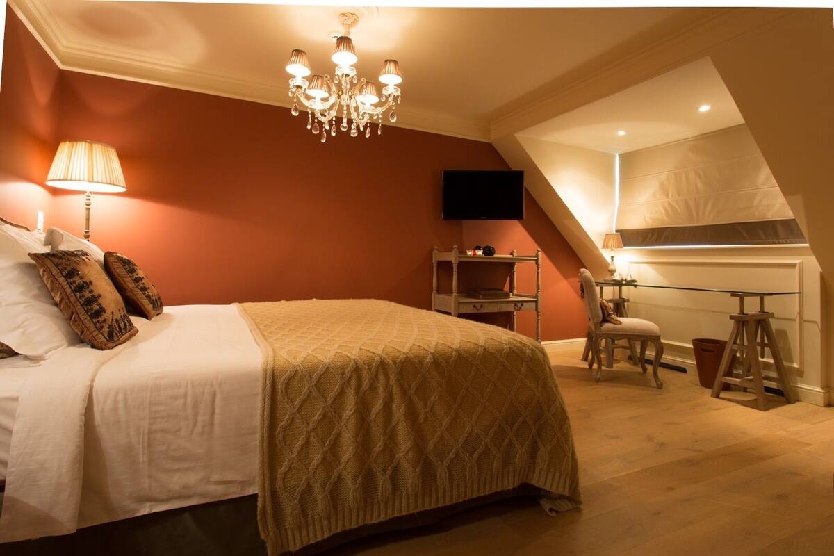 A bedroom with a large bed, a chandelier, a desk with a chair, and a TV on the wall.