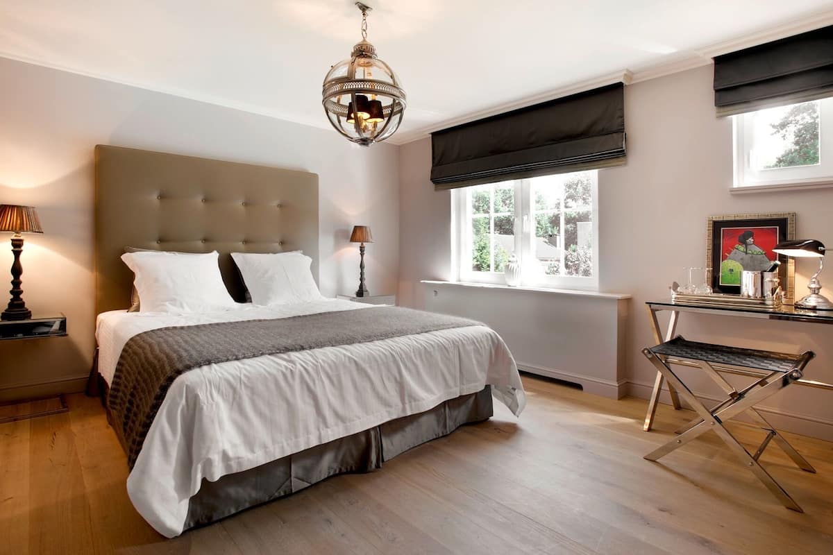 A bedroom with a large bed, a desk with a chair, a window with a view, and a hanging light fixture.