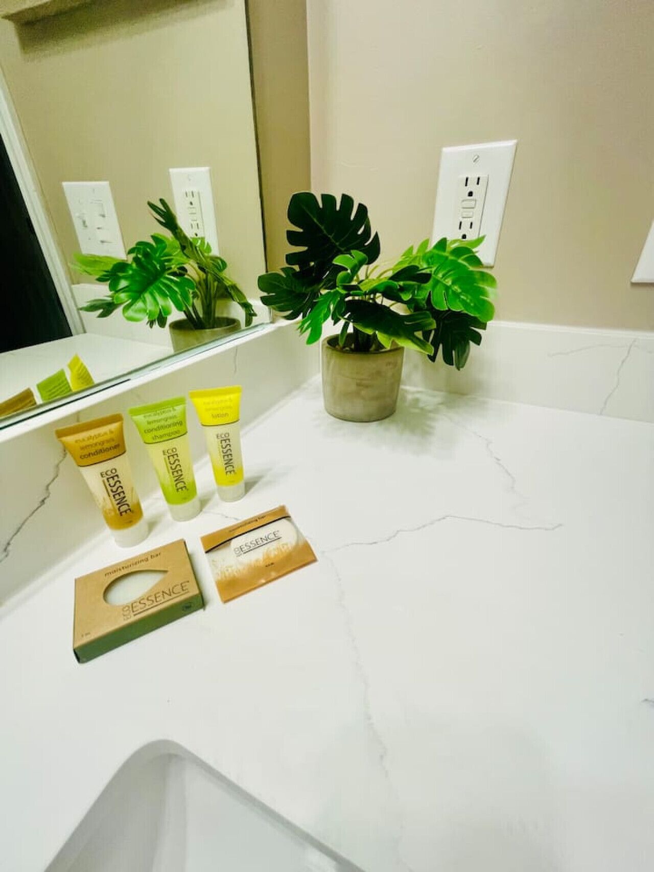 A bathroom with a white marble countertop, a potted plant, and three bottles of ESSENCE body lotion.