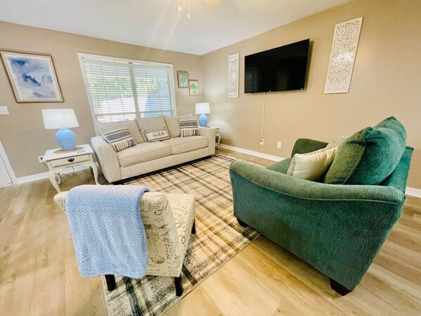 A living room with a beige sofa, a green armchair, a plaid rug, and a flat-screen TV mounted on the wall.