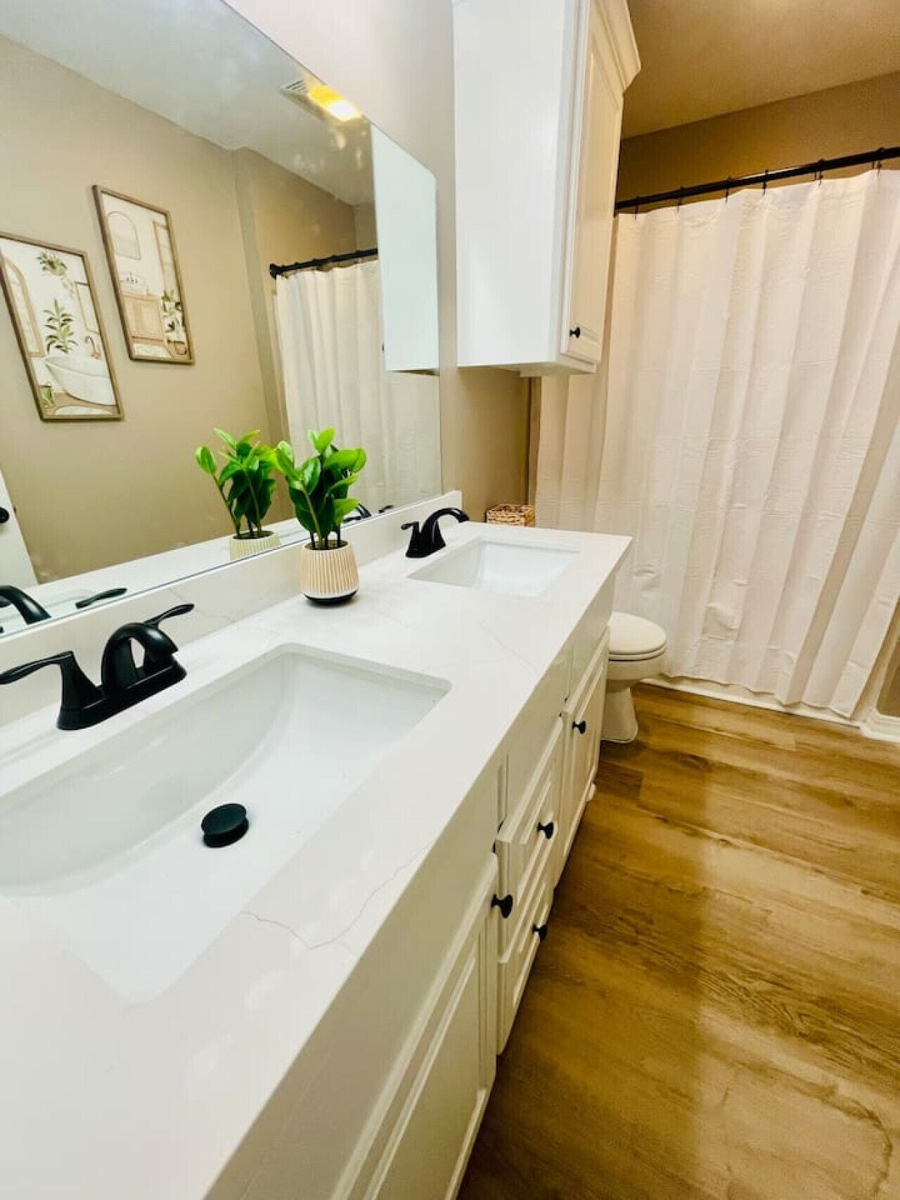 A bathroom with a white sink, two black faucets, a mirror, and a toilet. There are two potted plants on the counter.