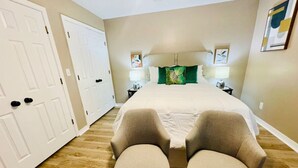 A bedroom with a bed, two chairs, a nightstand with a lamp, and a door.