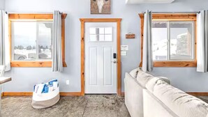 Una habitación con una puerta blanca, dos ventanas con marcos de madera, un sofá color beige y un cuadro de una cabeza de toro en la pared.