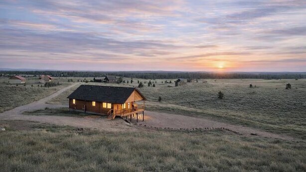 Una cabaña de madera con un porche, rodeada de campos abiertos y una puesta de sol al fondo.
