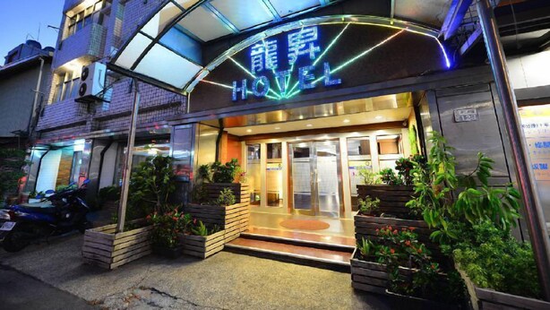 Een hotelentree met een neonreclame, glazen deuren en een klein terras met planten.