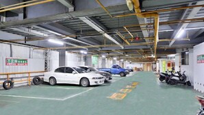 Een parkeergarage met meerdere auto's en een scooter, industriële luifel met blootliggende leidingen, en een groene vloer met pijlen voor de richting.