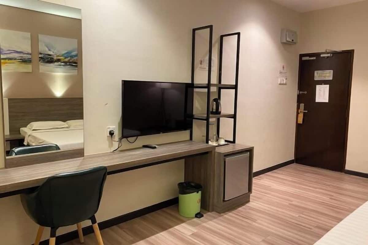A hotel room with a bed, a desk with a computer, a TV, a mirror, a small cabinet, and a trash bin.