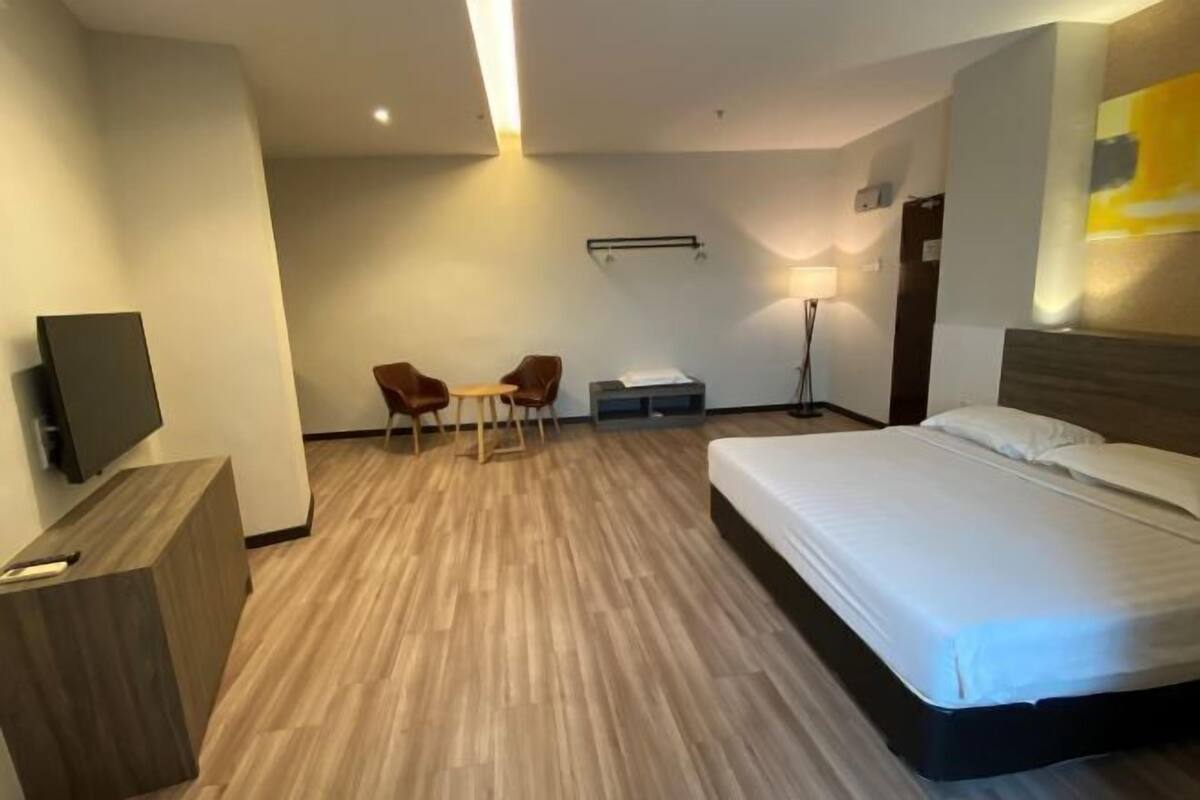 A hotel room with a bed, a desk, two chairs, a TV, a lamp, and a wall-mounted air conditioning unit.