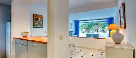 A kitchen with white cabinets, a wooden countertop, and a bowl of fruit. There is a window with blue curtains and a view of greenery outside.